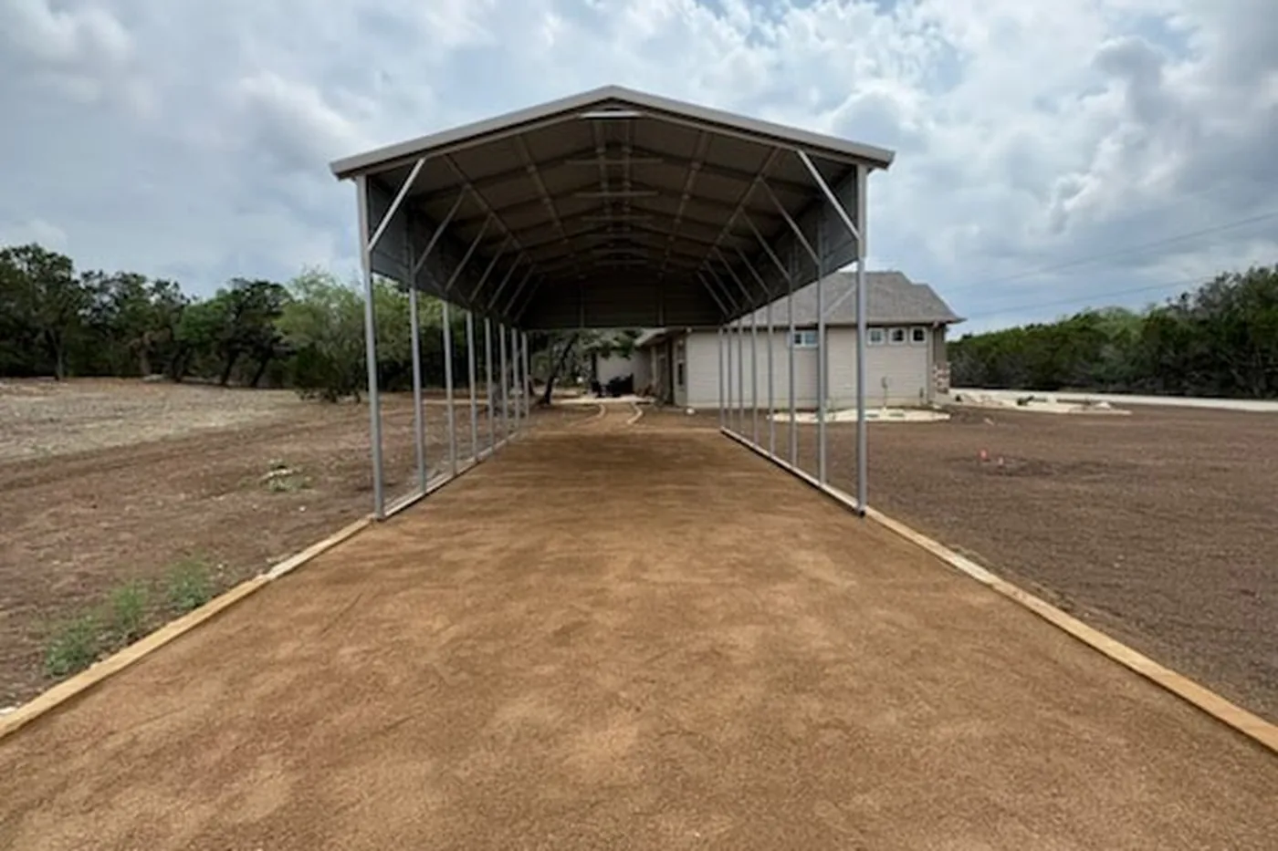 Graded caliche driveway running under a metal carport to a Hill Country home